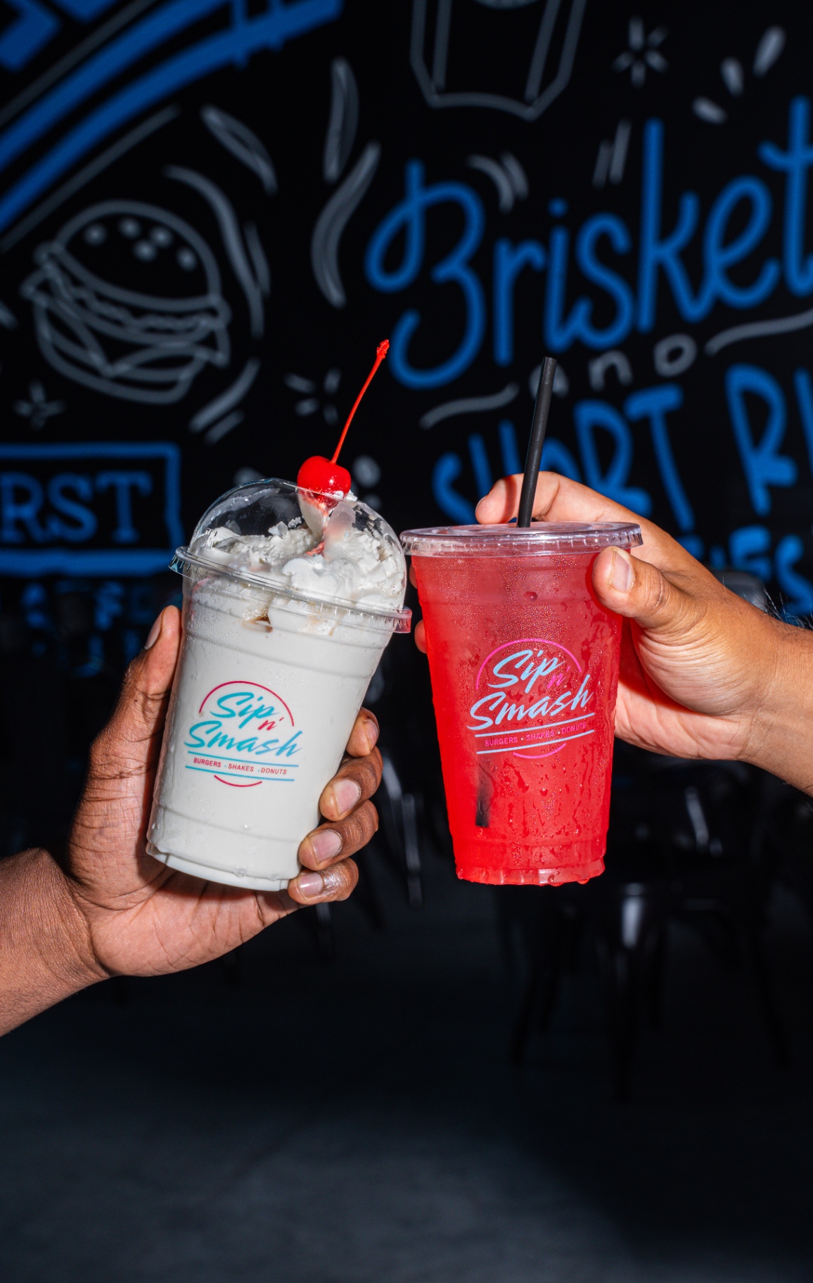 Two hands holding Sip N Smash shake and lemonade cups in front of brisket and short rib mural